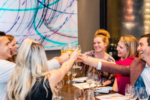A group of friends cheers inside of Burgundy Swine, a local wine bar in the Victory Park neighborhood of Dallas, Texas. The atmosphere is bright, modern and inviting with artwork surrounding the walls.